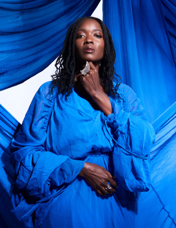 Une femme se tient debout, très droite dans une robe bleu. derrière elle, un tissus du même bleu forme des rideaux