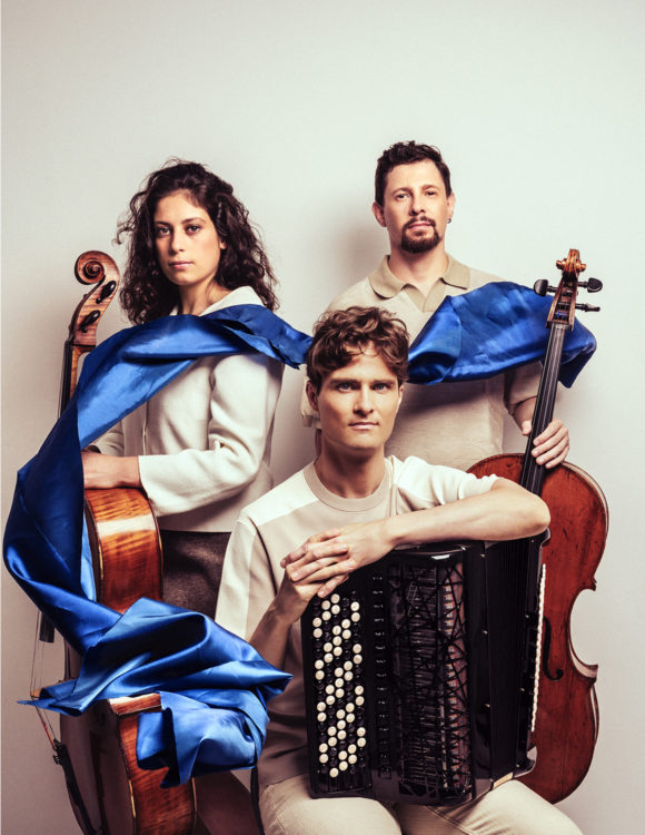 Une femme et 2 hommes posent avec leurs instruments, un accordéon et 2 violencelles. Un ruban bleu passe les relie.