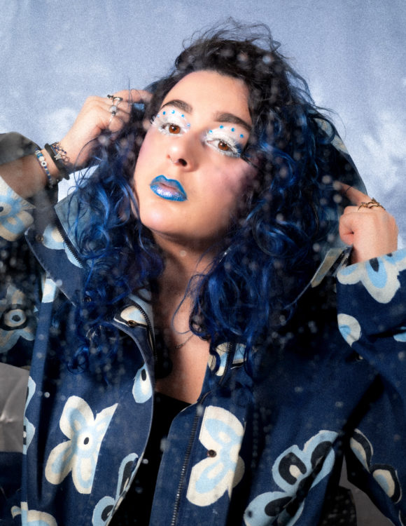 Une femme maquilée de façon très originale avec des cils longs et blancs et un rouge à lèvre bleu. Un vêtement ample; bleu marine à grosses fleurs blanches.