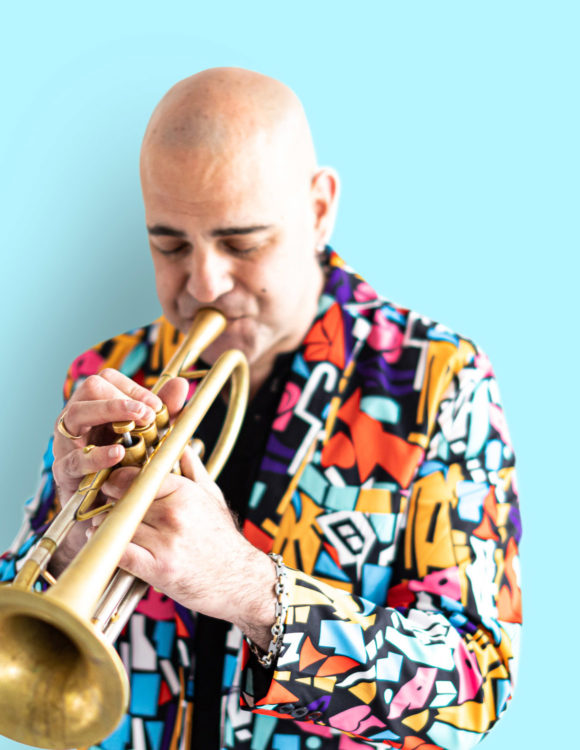 Un musicien souffle dans sa trompette, veste très colorée, le mur du fond est bleu ciel
