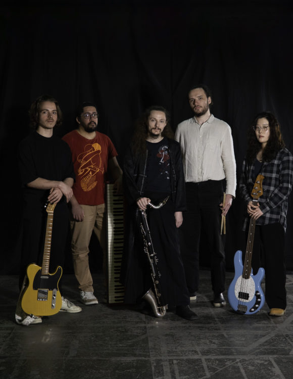 1 musicienne et 4 musiciens se tiennent debout, alignés devant un mur noir. Leurs instruments sont posés à terre devant eux.